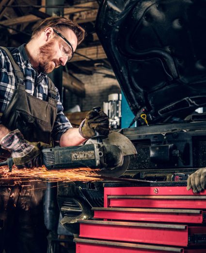 Bearded mechanic cuts steel car part with an angle grinder in a garage.